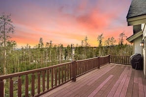 Una terraza de madera con barandilla, con vistas a un área boscosa y un atardecer.