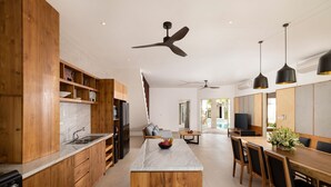 A modern kitchen with wooden cabinets, a marble island, and a dining area with a long table and chairs. - Villa Christie by Nakula (Seminyak)