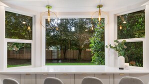 A kitchen with a large window overlooking a garden, three bar stools, and a white countertop.