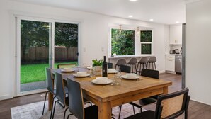 A dining area with a wooden table, chairs, and a chandelier. There is a kitchen in the background with white cabinets and a dishwasher.