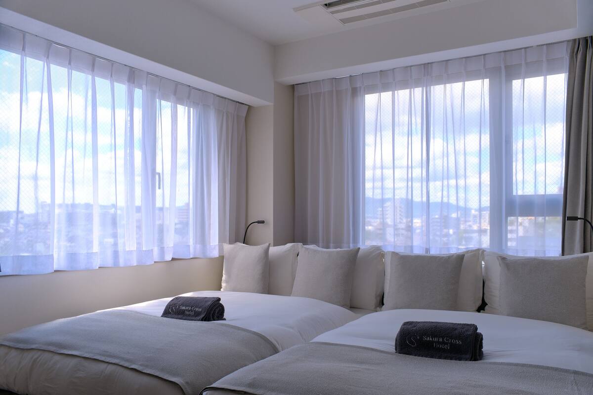 A hotel room with two beds, white bedding, and a view of the city through sheer curtains.