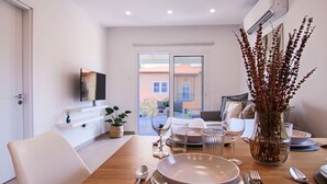 Un coin repas avec une table en bois dressée pour quatre personnes, un vase contenant des plantes séchées et une vue sur l’extérieur grâce à une porte vitrée.