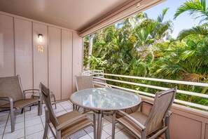 A balcony with a glass table and four chairs, surrounded by a railing and lush greenery.