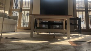Ein Wohnzimmer mit einem großen Fernseher auf einem Holztisch, einer Couch und einem gemusterten Teppich. Große Fenster mit Gitterstäben bieten Blick auf die Gebäude draußen.