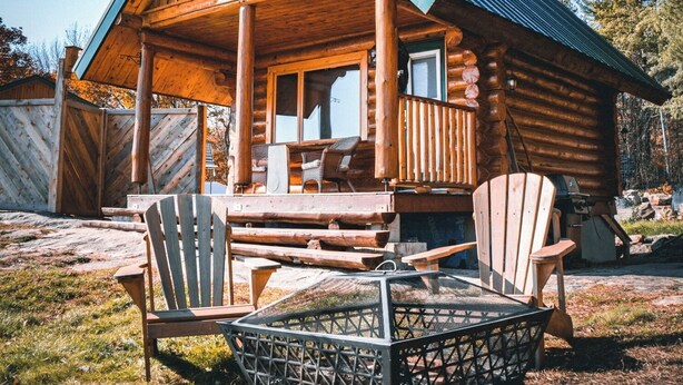 A wooden log cabin with a porch, two Adirondack chairs, and a fire pit.