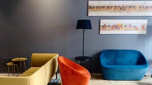 Una sala de estar moderna con un sofá amarillo, un sillón naranja y un sofá azul. Hay una alfombra estampada en el suelo, una lámpara de pie negra y dos cuadros en la pared.
