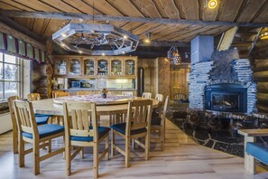 A rustic cabin interior with a wooden dining table, chairs, a stone fireplace, and a kitchen area with wooden cabinets and a hanging light fixture. - Rukan Salonki Chalets (Kuusamo)