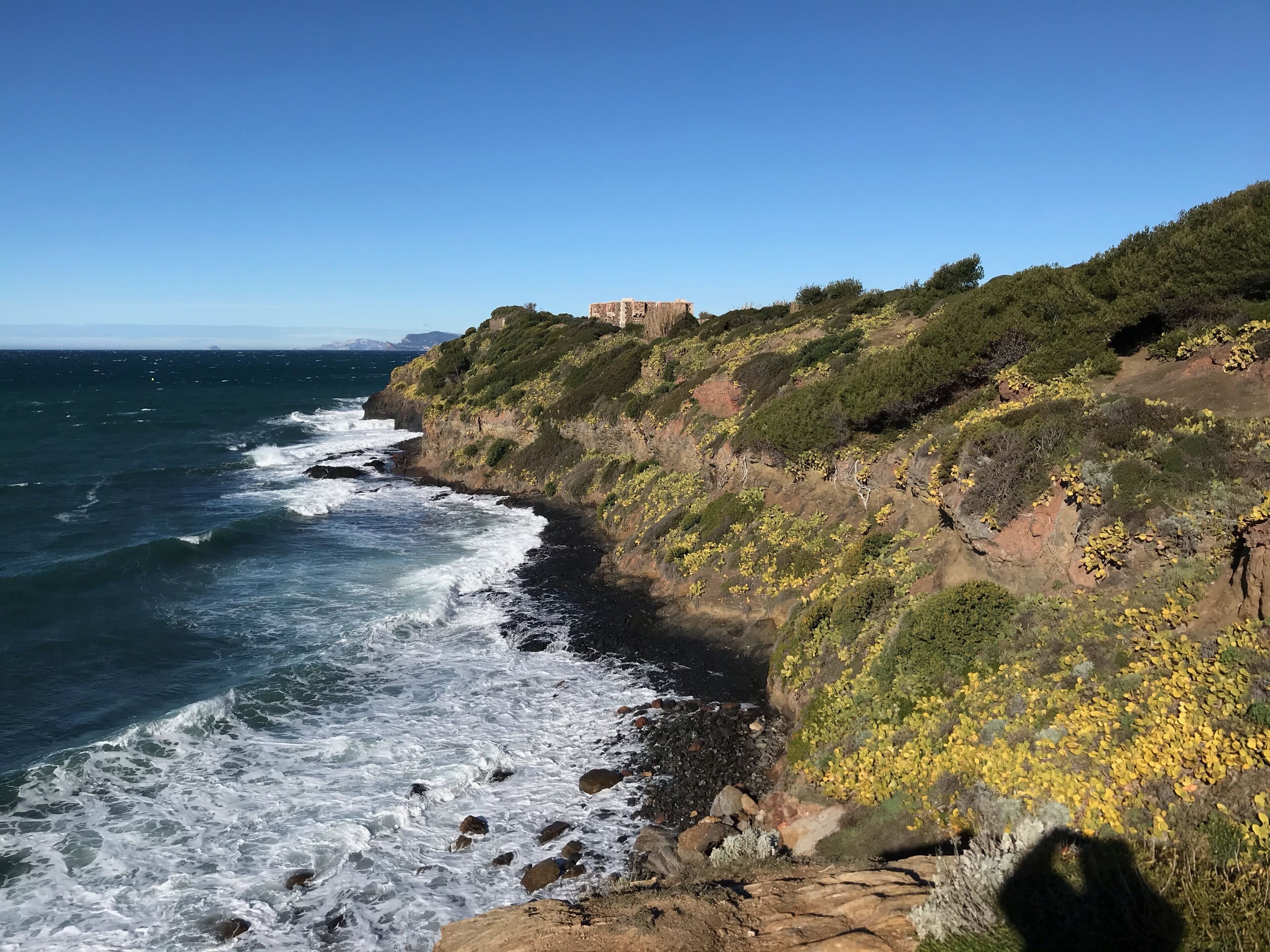 A coastal landscape with rocky cliffs, waves crashing against the shore, and a building on the hillside.