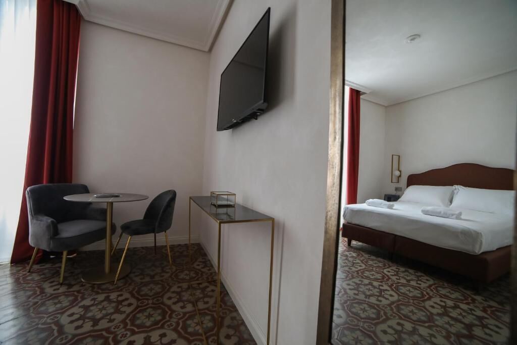 A hotel room with a bed, a TV mounted on the wall, a small table with chairs, and a patterned carpet.