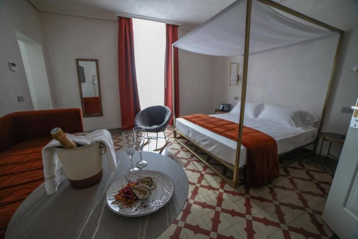 A hotel room with a bed, a chair, a table with a bottle and glasses, and a checkered floor.