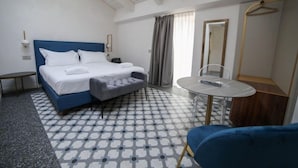 A hotel room with a bed, a small table, a chair, a mirror, and a hanging coat rack.