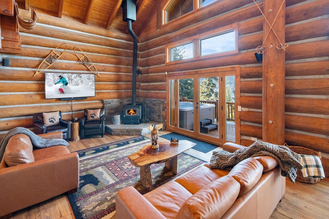 Interno di una baita di legno con camino, divani in pelle, televisore e una grande finestra.