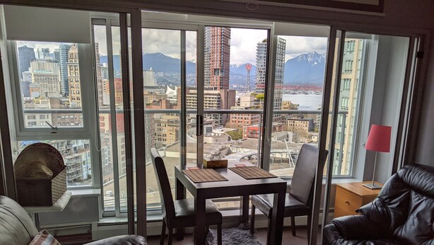 A modern living room with a large window offering a city view, a dining table with chairs, and a leather sofa. - Ocean, Mountain, City Views, Fully Furnished, Downtown Vancouver (Vancouver)