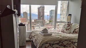 A bedroom with a large window offering a city view, a neatly made bed with a floral bedspread, and a desk with a lamp. - Ocean, Mountain, City Views, Fully Furnished, Downtown Vancouver (Vancouver)