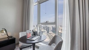 Una habitación de hotel moderna con un ventanal grande, una mesa redonda, dos sillas, una computadora portátil y un adorno en una mesita de noche.