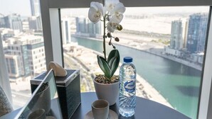 Una mesa con una orquídea en maceta, una botella de agua y una taza, con vista a un paisaje urbano a través de una ventana.