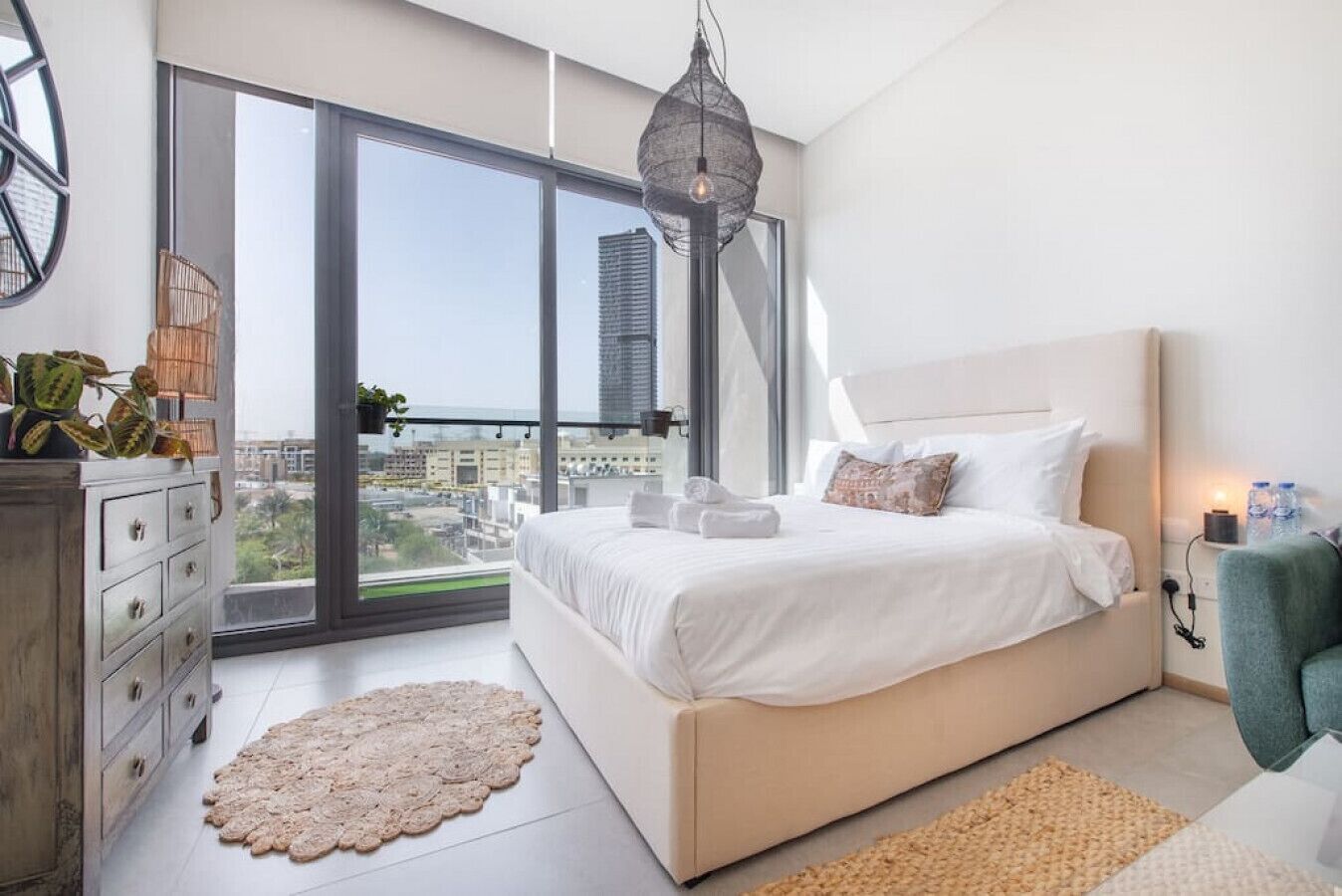 Une chambre moderne avec un grand lit, une commode, un tapis rond et une vue sur la ville grâce à de grandes fenêtres.