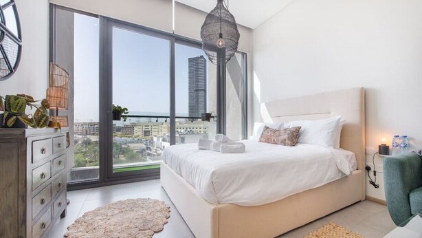 A modern bedroom with a large bed, a dresser, a round rug, and a view of the city through large windows.