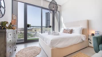 A modern bedroom with a large bed, a dresser, a round rug, and a view of the city through large windows.