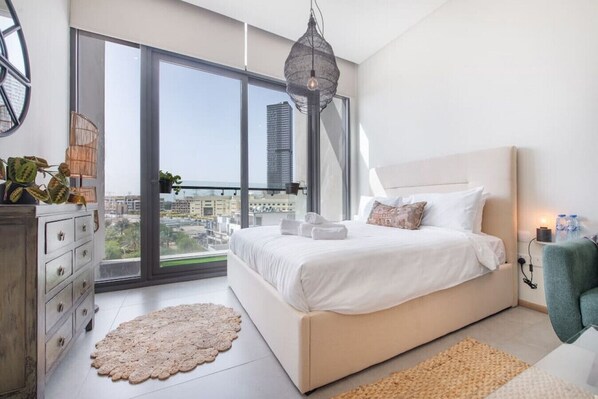 A modern bedroom with a large bed, a dresser, a round rug, and a view of the city through large windows.