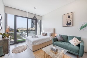 A modern bedroom with a large bed, a green sofa, and a city view through floor-to-ceiling windows.