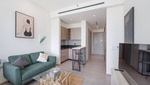 A modern apartment with a green sofa, a kitchen with wooden cabinets, a dining area with a bar stool, and a flat-screen TV.