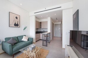 A modern apartment with a green sofa, a kitchen with wooden cabinets, a dining area with a bar stool, and a flat-screen TV.