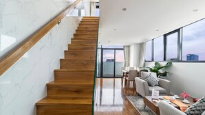 Un interior moderno con una escalera de madera, un comedor con una mesa dispuesta para dos personas, y ventanas amplias que ofrecen vistas del horizonte urbano.