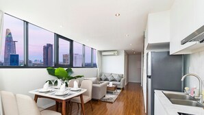 Un apartamento moderno con comedor, sala y cocina. Cuenta con ventanas amplias con vista a la ciudad, una mesa de comedor para cuatro personas, un sofá y una cocina con refrigerador y lavaplatos.