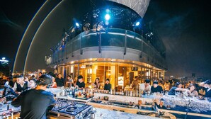 Un bar concurrido con cabina de DJ, gente socializando y una vista panorámica de la ciudad al fondo.