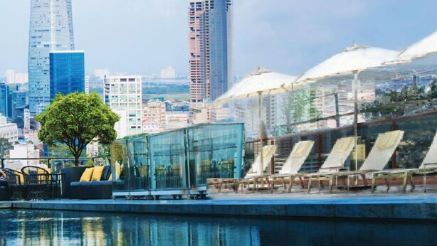 Un moderno paseo marítimo urbano con piscina, sillas de descanso y un edificio de vidrio con un árbol en primer plano.