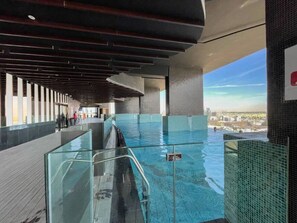 A modern indoor swimming pool with clear glass walls, a wooden deck, and a view of the cityscape.