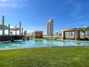 A swimming pool with a grassy area, a pavilion, and a tall building in the background.