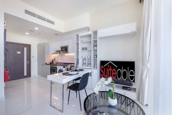 A modern kitchen with white cabinets, a microwave, and a dining area with a white table and black chairs. There is a TV mounted on the wall with a 'suiteable' sign above it.