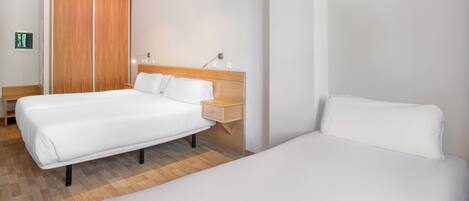 A hotel room with two single beds, a wooden wardrobe, a bedside table with a lamp, and a small bench.