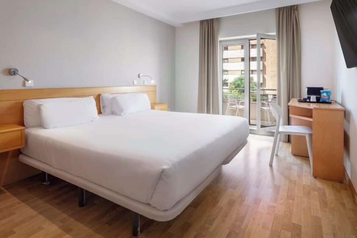 Una habitación de hotel con una cama grande, un escritorio y un balcón con vistas a edificios.