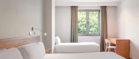 A hotel room with two beds, a desk, a chair, a window with curtains, and a bedside lamp.
