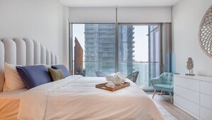 Ein modernes Schlafzimmer mit einem großen Bett, Blick auf hohe Gebäude und einem Balkon mit einem Stuhl.