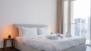 Un lit bien fait, avec une literie blanche, une tête de lit grise et une table de chevet sur laquelle se trouve une lampe. Un oreiller avec le mot « Suiteable » est posé sur le lit.