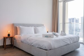 Un lit bien fait, avec une literie blanche, une tête de lit grise et une table de chevet sur laquelle se trouve une lampe. Un oreiller avec le mot « Suiteable » est posé sur le lit.