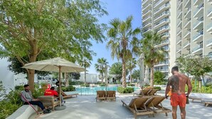 Un espace aménagé au bord de la piscine, avec des chaises longues, un palmier et un grand immeuble en arrière-plan.