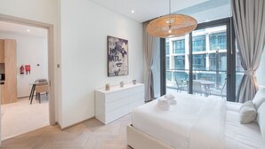 A modern bedroom with a large bed, a white dresser, a dining area with a table and chairs, and a view of the city through large windows. - JVC-Signature Livings 103 N · Modern flat with KING SIZE bed and HIGH SPEED Wifi (Dubai)