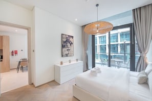 Un dormitorio moderno con una cama grande, un aparador blanco, un área de comedor con mesa y sillas, y vistas de la ciudad a través de amplias ventanas.