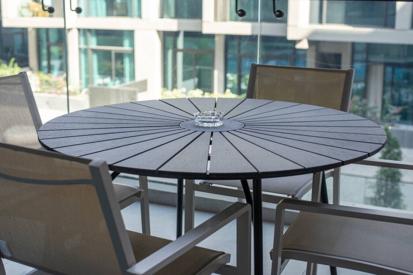 Une table ronde ornée d’un objet en verre au centre, entourée de chaises à structure métallique et à assise rembourrée, le tout installé sur un balcon offrant une vue sur des bâtiments et de la verdure.