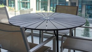 A round table with a central glass object, surrounded by chairs with metal frames and cushioned seats, set on a balcony with a view of buildings and greenery. - JVC-Signature Livings 103 N · Modern flat with KING SIZE bed and HIGH SPEED Wifi (Dubai)