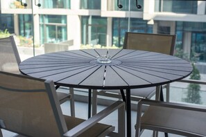 Une table ronde ornée d’un objet en verre au centre, entourée de chaises à structure métallique et à assise rembourrée, le tout installé sur un balcon offrant une vue sur des bâtiments et de la verdure.