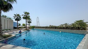 Ein Schwimmbecken mit Liegestühlen und Palmen, ein modernes Gebäude und ein Riesenrad im Hintergrund.