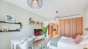 A modern bedroom with a bed, bedside table, and a desk with chairs. There is a kitchenette in the background.