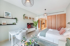 A modern bedroom with a bed, bedside table, and a desk with chairs. There is a kitchenette in the background.