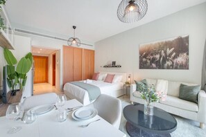 A modern hotel room with a bed, a sofa, a dining table set for two, and a large window.